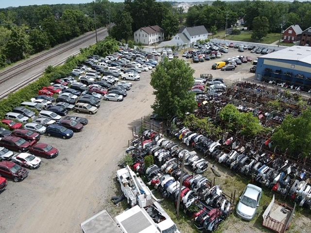 An aerial view of a vehicle salvage yard with rows of parked and dismantled cars near railroad tracks and a warehouse.