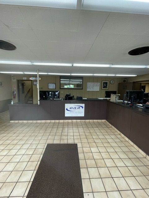 An office reception area with a dark brown counter, a logo, and tile floors under long, rectangular ceiling lights.
