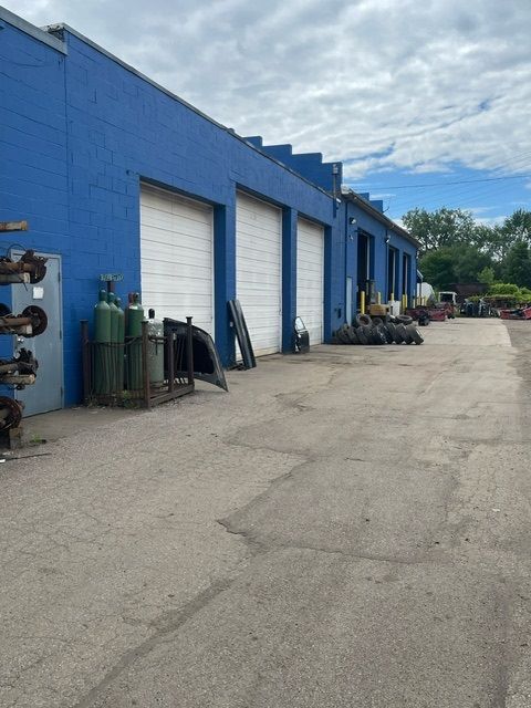 A bright blue industrial building with several white roll-up garage doors, gas cylinders, and tires in an outdoor lot.