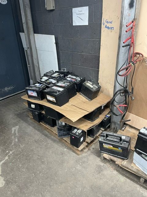 A stack of black lead-acid batteries arranged on pallets against a cinder block wall, with jumper cables hanging nearby.