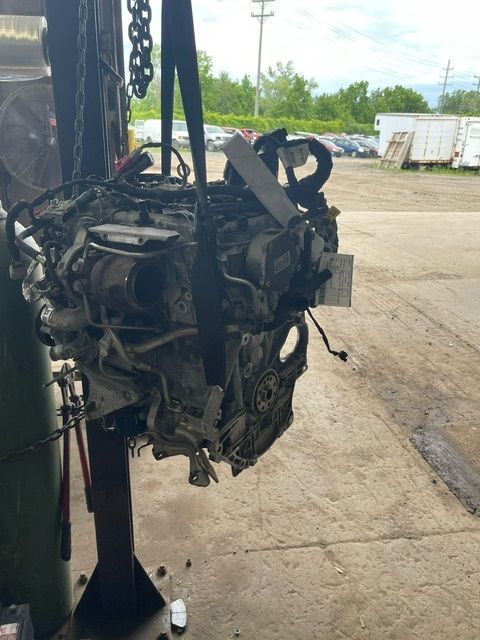 A car engine suspended by a black strap from a hoist in a repair shop or salvage yard.
