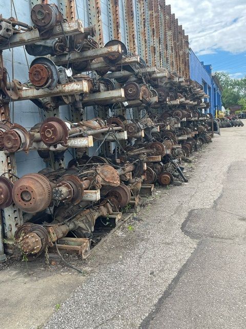 A large rack filled with salvaged automotive axles and drivetrain components stacked on a paved outdoor lot.