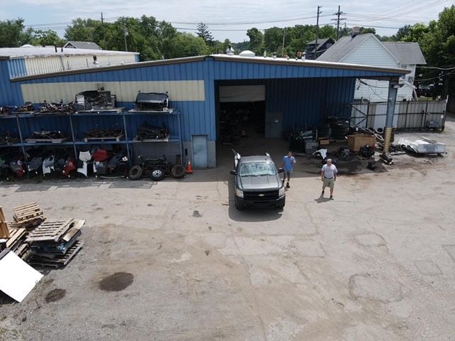 Two people stand near a dark pickup truck outside an automotive salvage warehouse with exterior parts shelving.