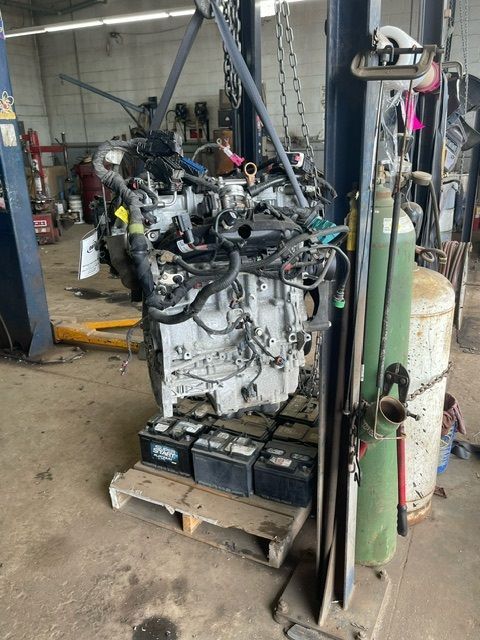 A car engine hangs from a crane above a wooden pallet loaded with several black lead-acid batteries in a repair garage.