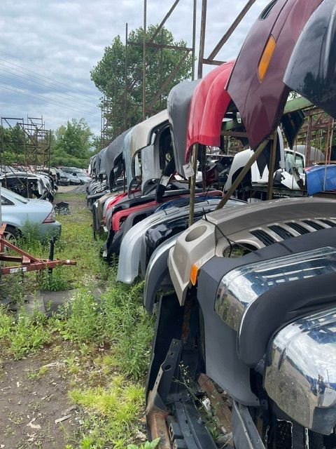 A row of assorted car bumpers and body panels hanging on an outdoor rack at a salvage yard under a cloudy sky.