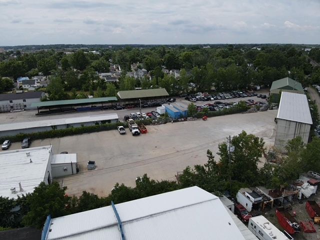 Aerial view of a paved commercial lot with parked vehicles, storage sheds, and metal structures under a cloudy sky.