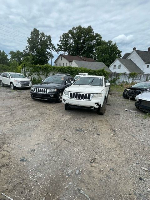Several vehicles, including two white and one black Jeep SUV, are parked in a gravel lot with houses in the background.