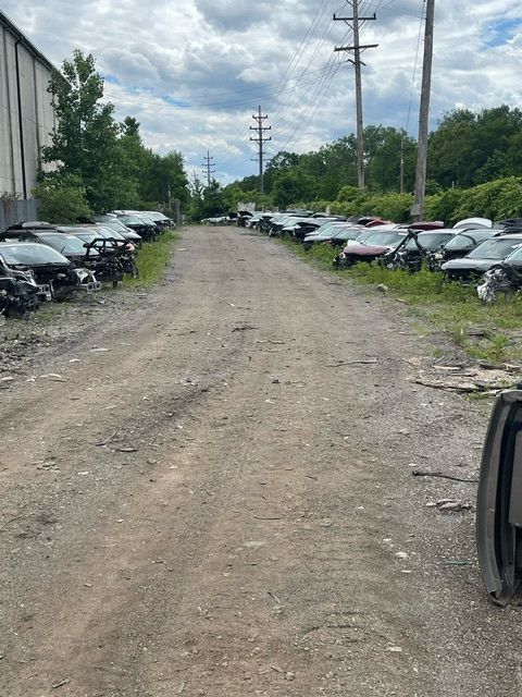 A gravel path winds through a salvage yard lined with rows of wrecked and dismantled cars under a cloudy sky.