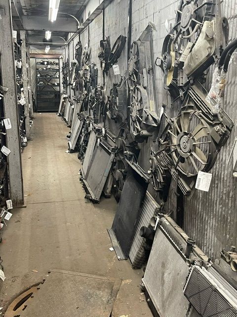 A narrow warehouse aisle lined with salvaged car radiators and cooling fans hanging on walls and placed on the floor.