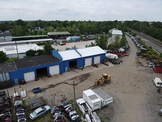 An aerial view of an industrial scrap yard with a blue workshop building, heavy machinery, and rows of salvaged vehicles.
