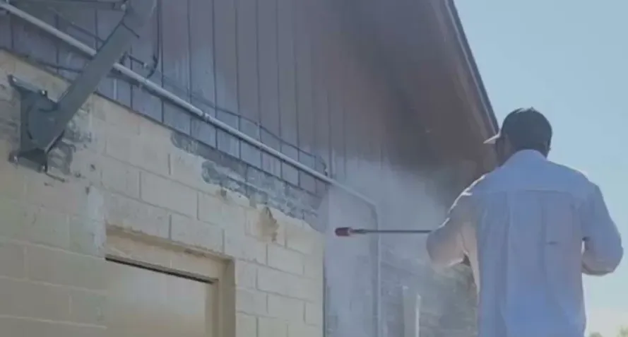 A person in a white shirt uses a pressure washer to clean a brick wall under a metal awning on a sunny day.