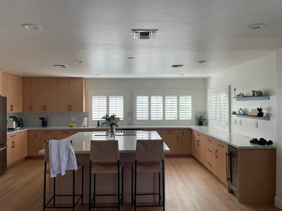 Modern kitchen with light wood cabinets, white countertops, an island with bar stools, and large windows with shutters.