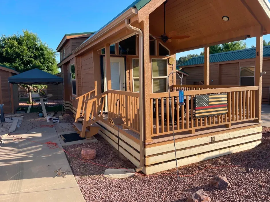A small, tan wood cabin with a covered front porch featuring a hanging American flag, set on a gravel lot under blue skies.