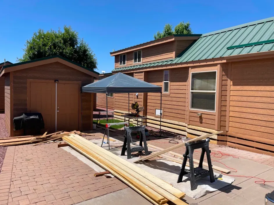 A construction project on a patio featuring wooden planks, two sawhorses, and a small gazebo next to a brown house.