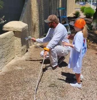A person in work clothes uses a paint sprayer on a stucco wall while a child stands nearby and watches.