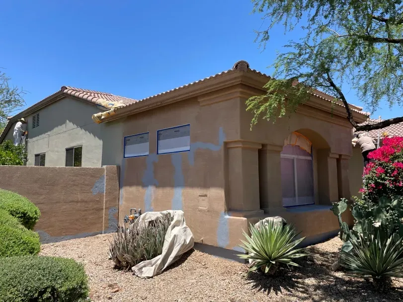 A worker on a ladder painting a tan stucco home with desert landscaping and a matching wall under a clear blue sky.