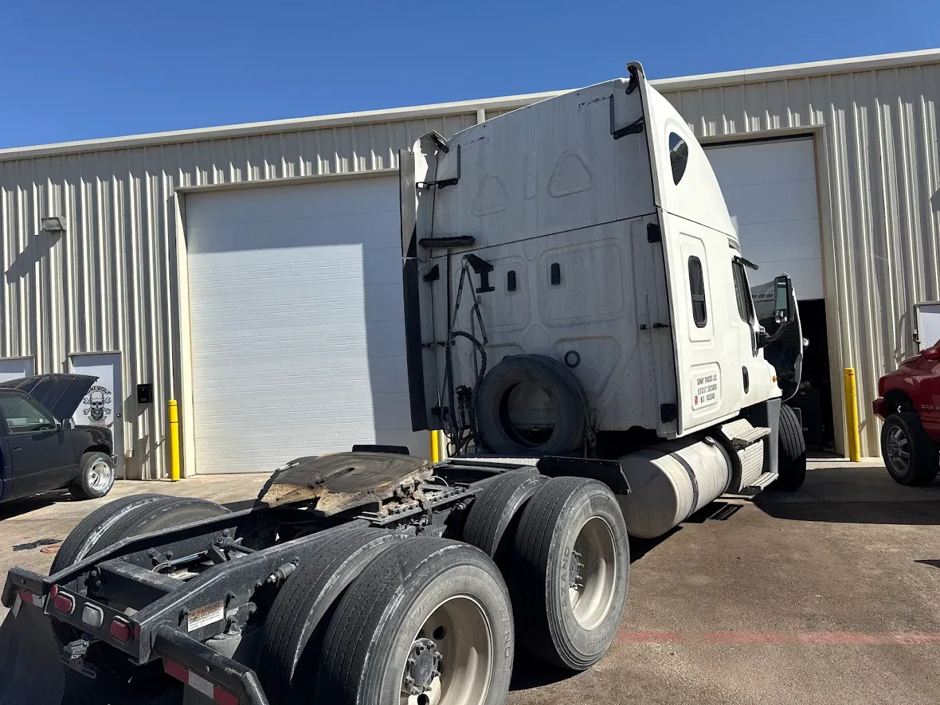 White semi-truck parked outside a building with garage doors, viewed from the rear, sunny day.