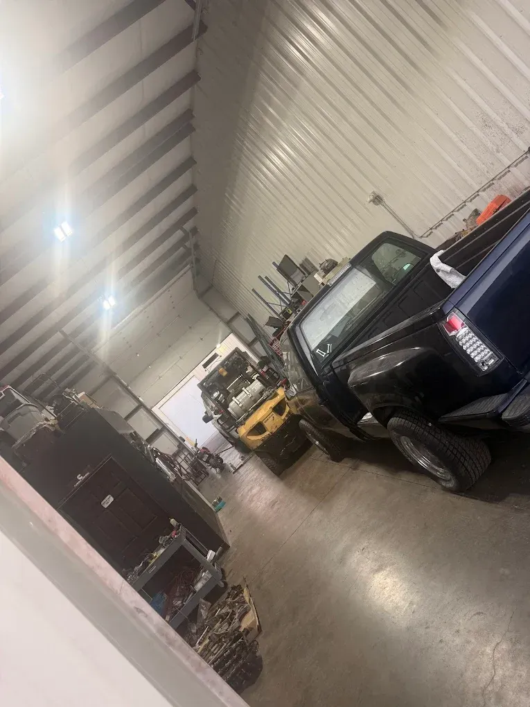 Inside a workshop, two trucks parked near a yellow forklift. Parts and shelves line the walls.
