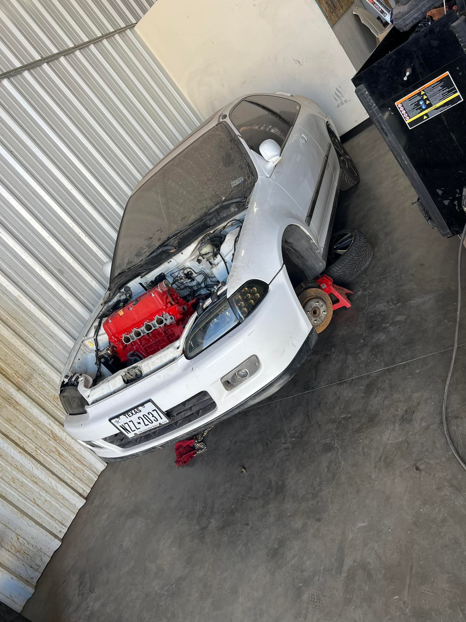 White car with red engine, hood open, on jack stands in front of a building.