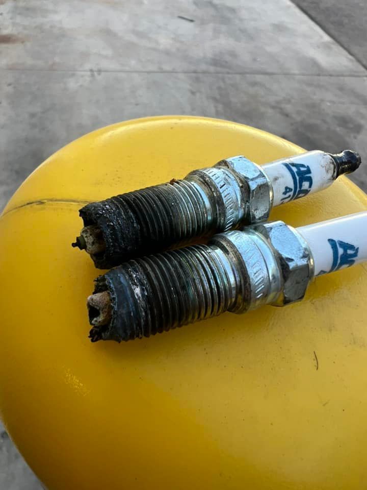 Two spark plugs, heavily fouled with black deposits, rest on a yellow surface.
