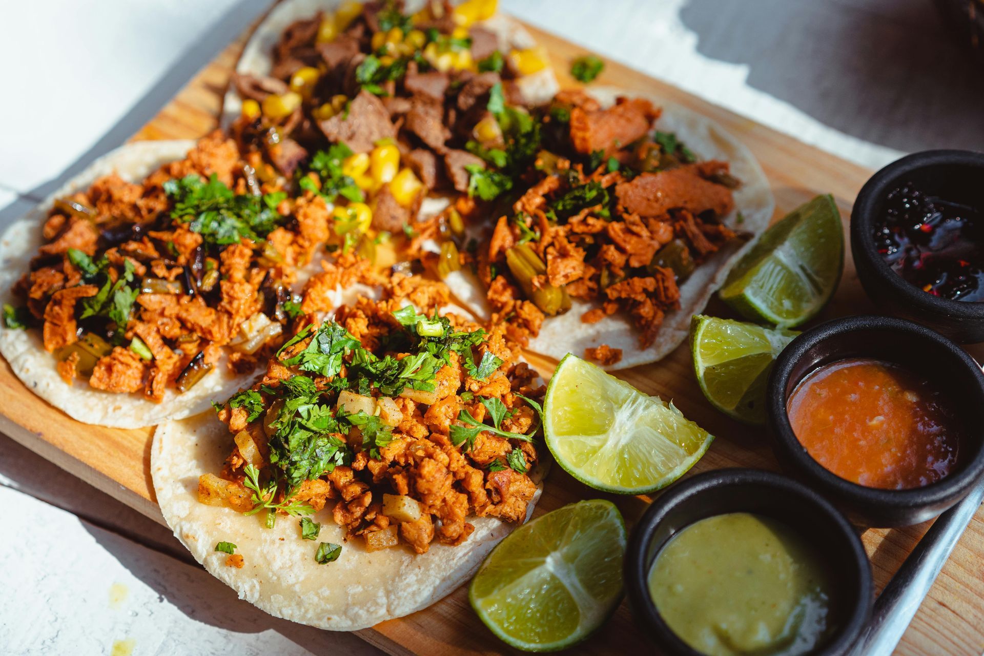 Tacos on a wooden board with toppings, limes, and dips.