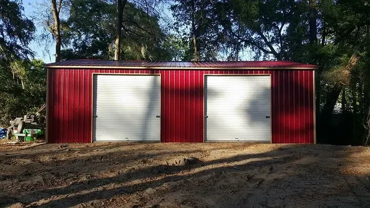 A red garage with two white garage doors is sitting in the middle of a dirt field.