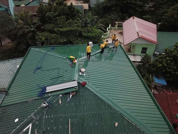 A group of workers in yellow shirts repairing the green, sloped metal roof of a building from an elevated view.