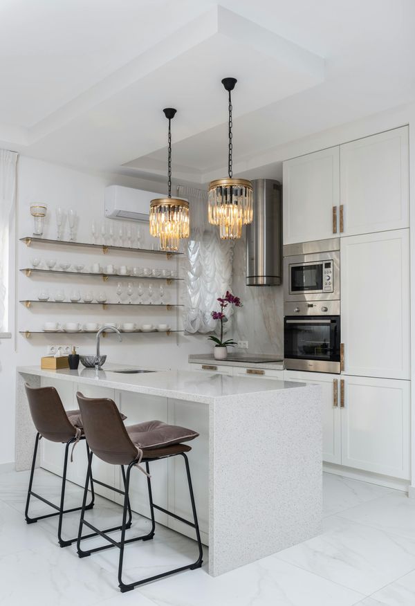 Modern white kitchen with a quartz island, two brown bar stools, gold pendant lights, and open shelving with glassware.