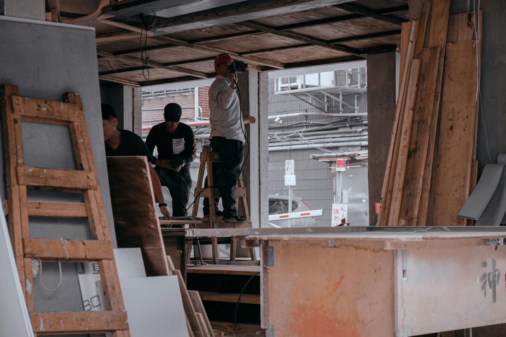 Construction workers work inside a building with wooden frames, scaffolding, and stacked materials in an open workspace.