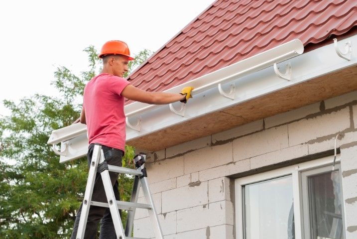 Worker on a ladder painting house eaves beside a red roof with a paint roller