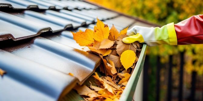 Gloved hand clearing autumn leaves from a gutter on a house roof