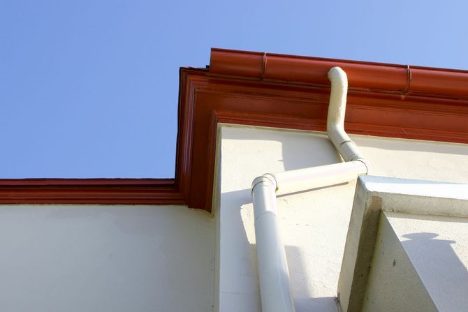 White building corner with red roof trim and a downspout against a blue sky