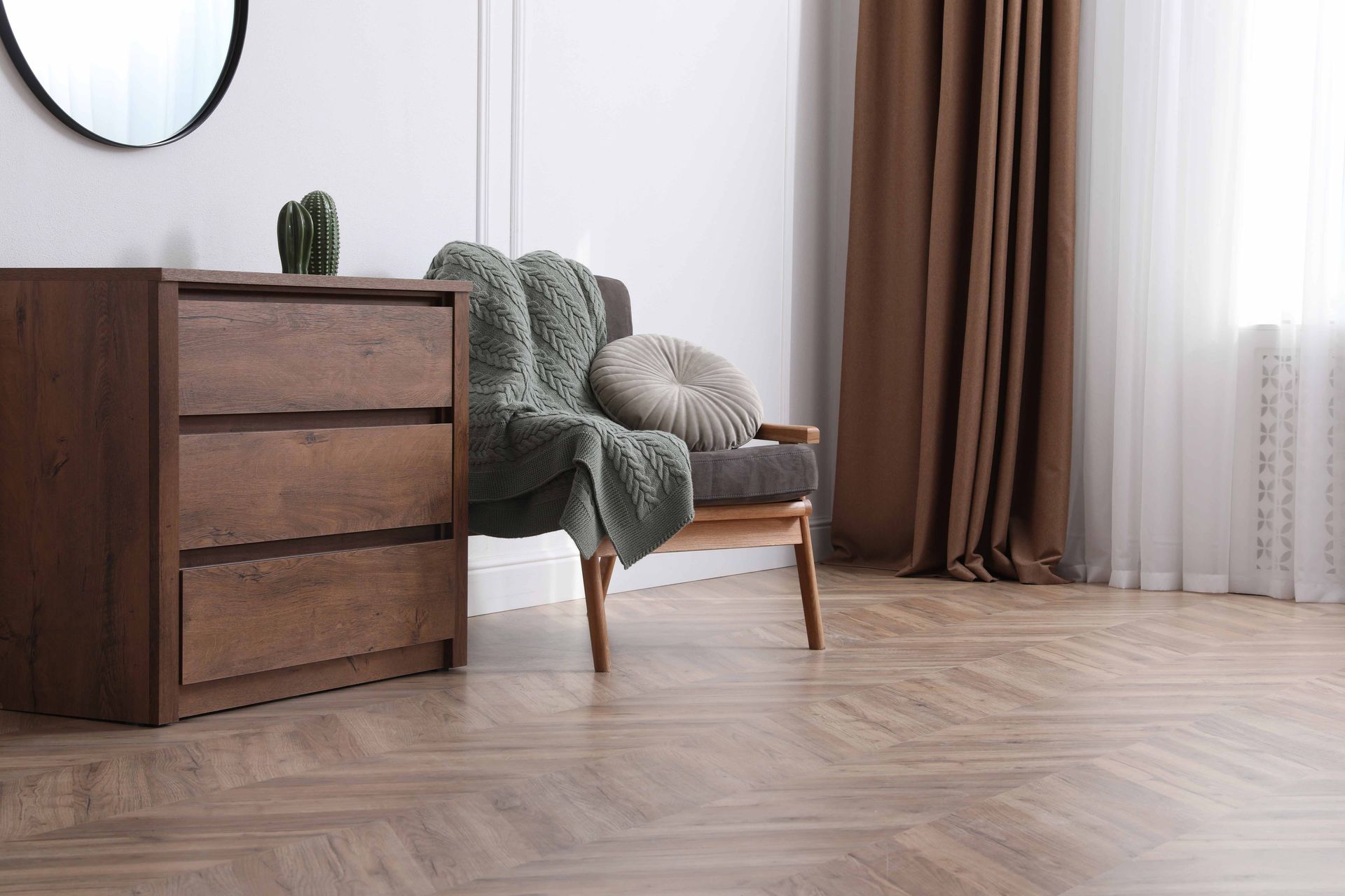 Wooden dresser, armchair with blanket and pillow, brown curtains, and a round mirror on a white wall.
