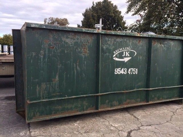 A large green JK Recycling skip bin in the centre of a parking lot, surrounded by pavement. A large green JK Recycling skip bin in the centre of a parking lot, surrounded by pavement.