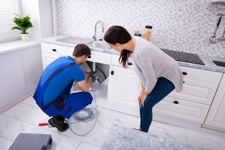 Plumber repairing a kitchen sink while another person watches in a white kitchen