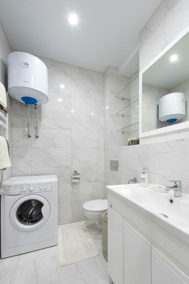 Small white bathroom with washer, toilet, sink vanity, mirror, and wall-mounted water heater.