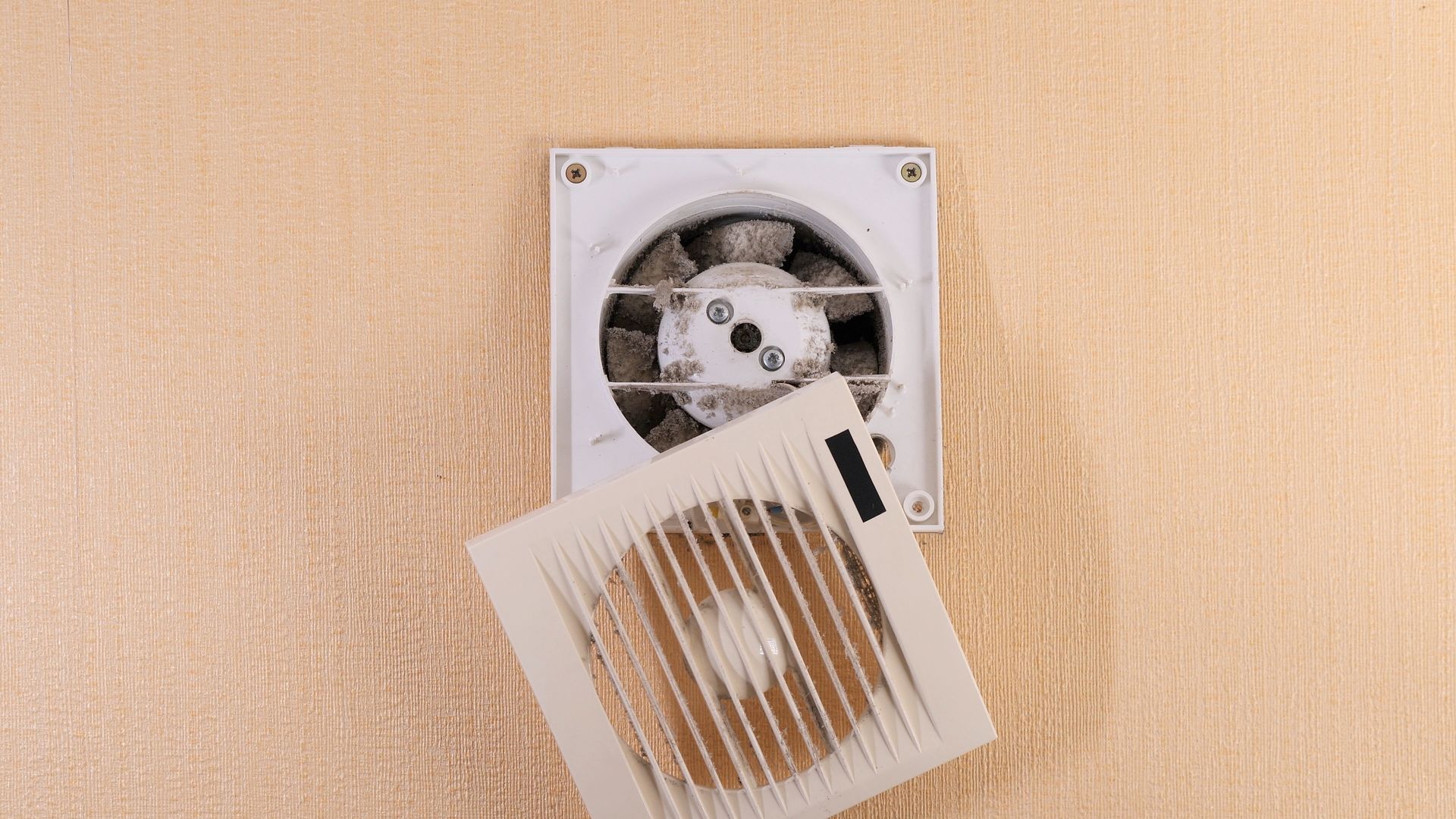 Damaged wall vent with detached grille hanging open against a beige wall