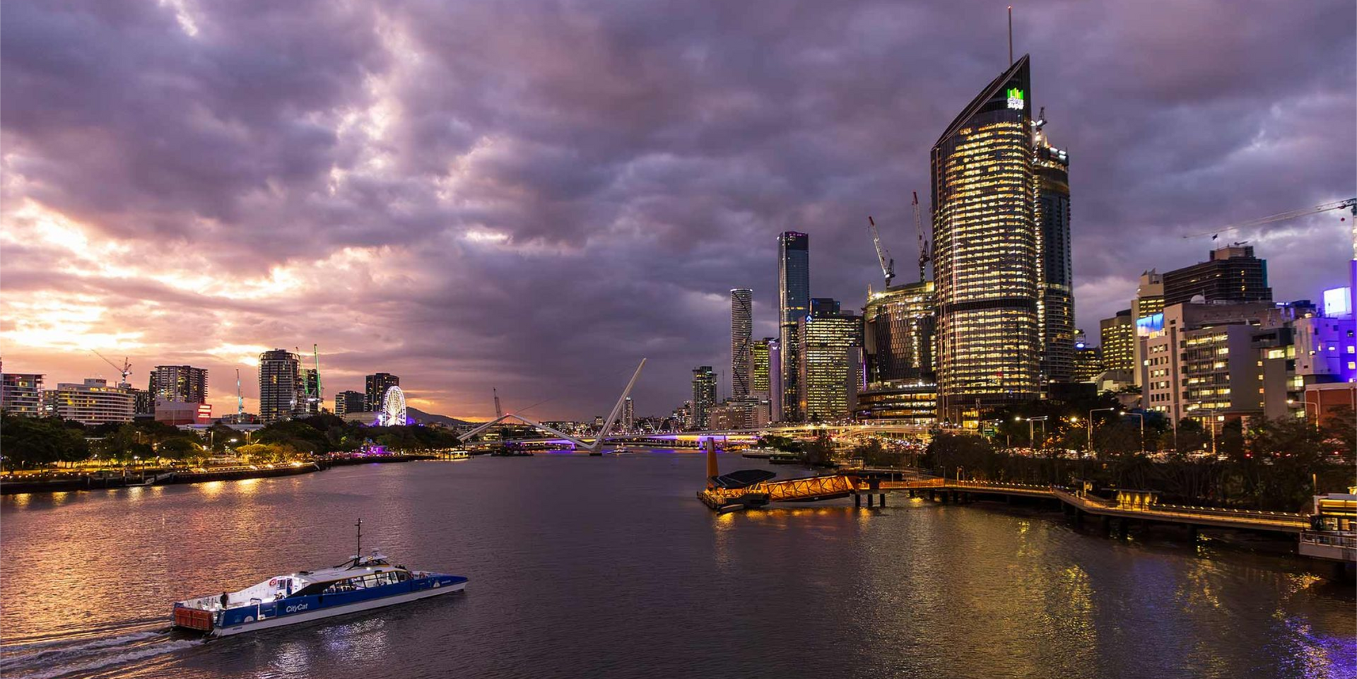 The Brisbane city skyline at dusk, seen across the river with a CityCat passing