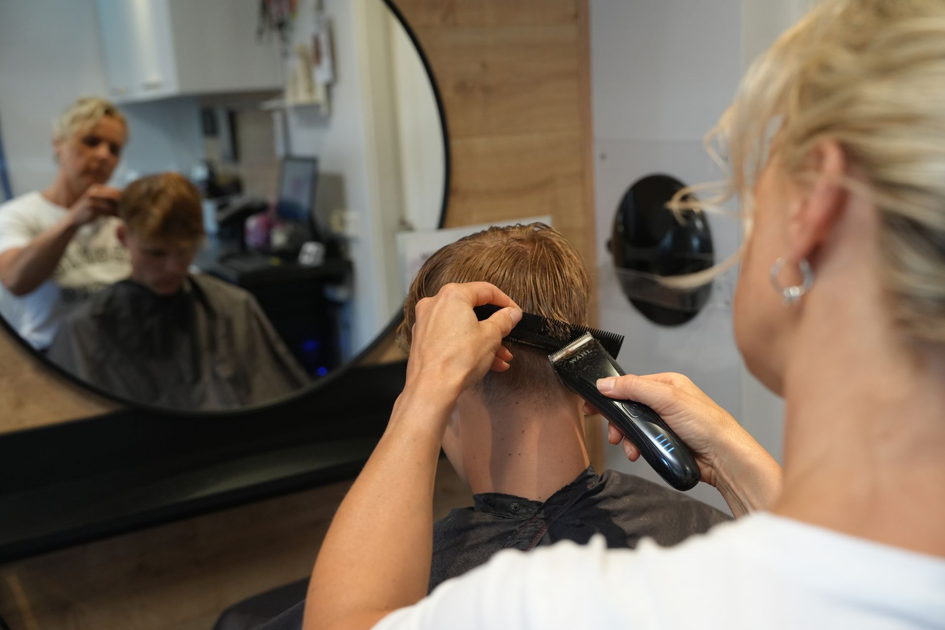 Een kapper knipt het haar van een klant met een tondeuse in een salon, zoals te zien is in een spiegel.