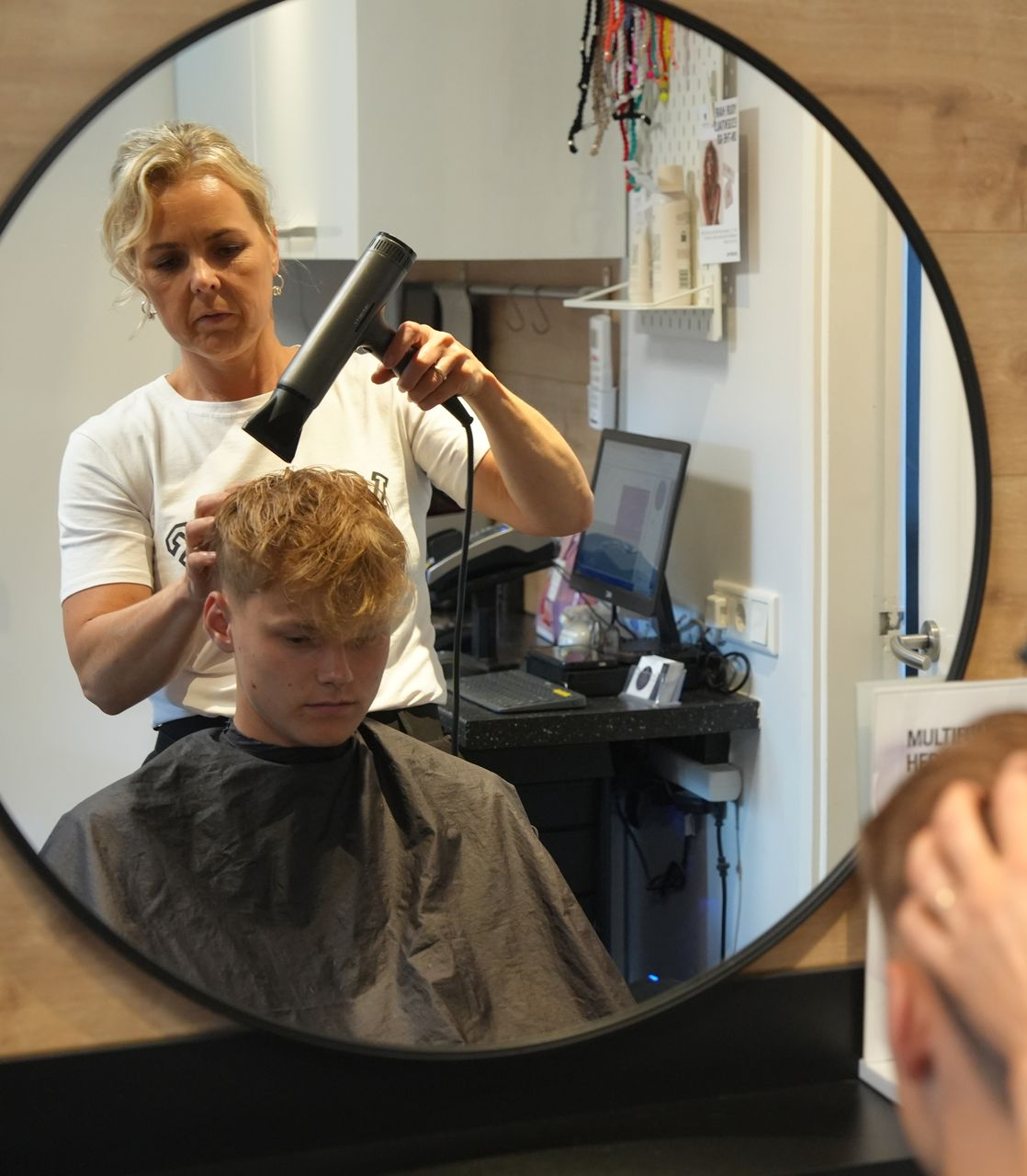 Een kapper föhnt het haar van een klant in een salon, gezien in een ronde spiegel.
