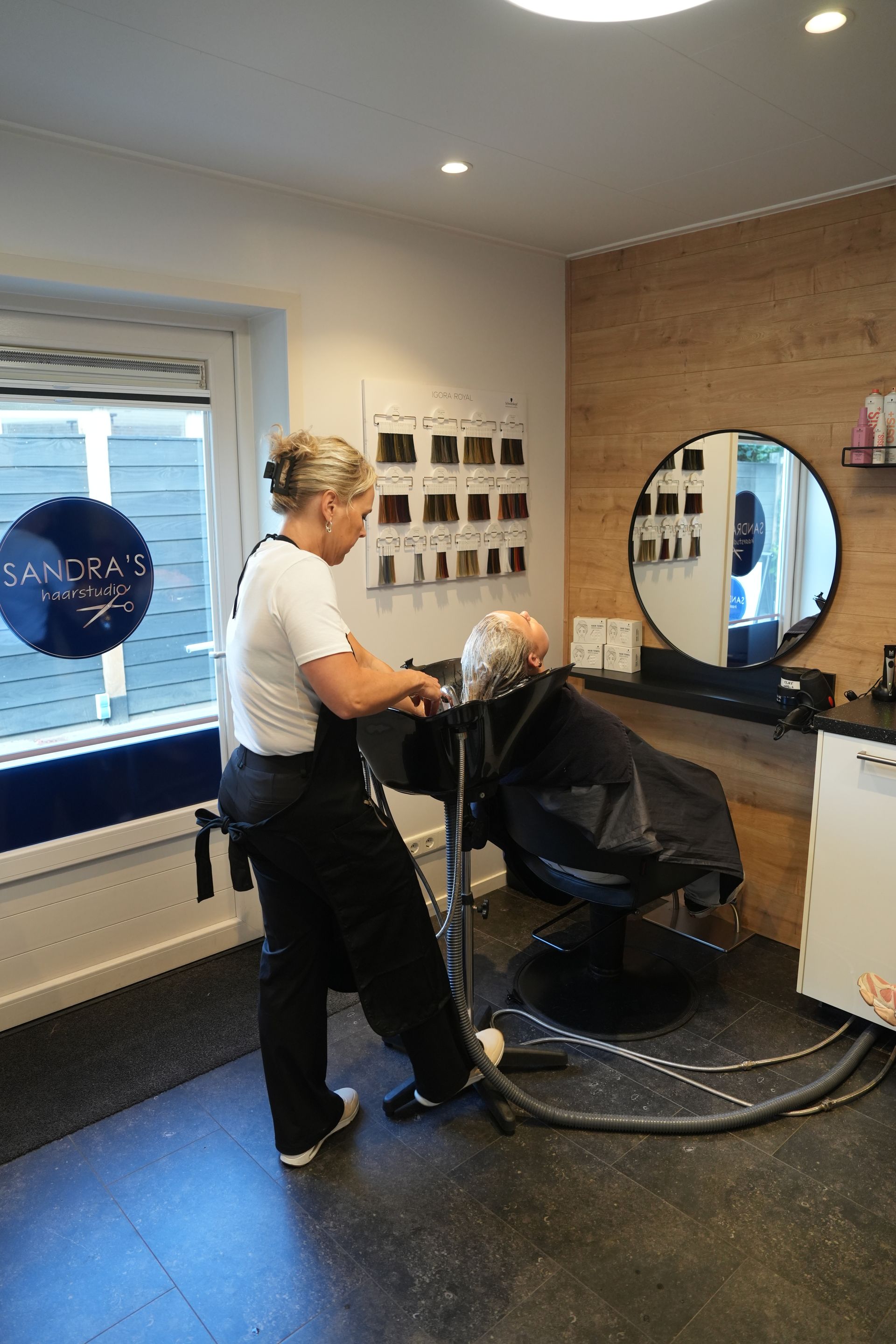 Een stylist föhnt het haar van een klant in een moderne salon met een ronde spiegel en een bakstenen accentmuur.