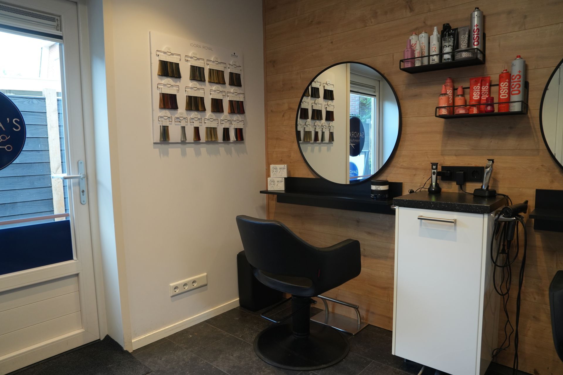 Salontafel met zwarte kappersstoel, ronde spiegel, planken met haarproducten en een witte kast.