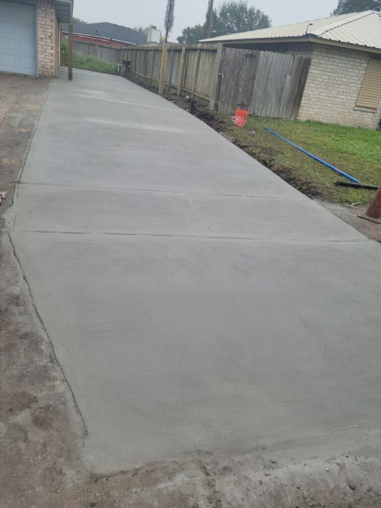 Newly poured concrete driveway extending from a building, beside a fence and grass.