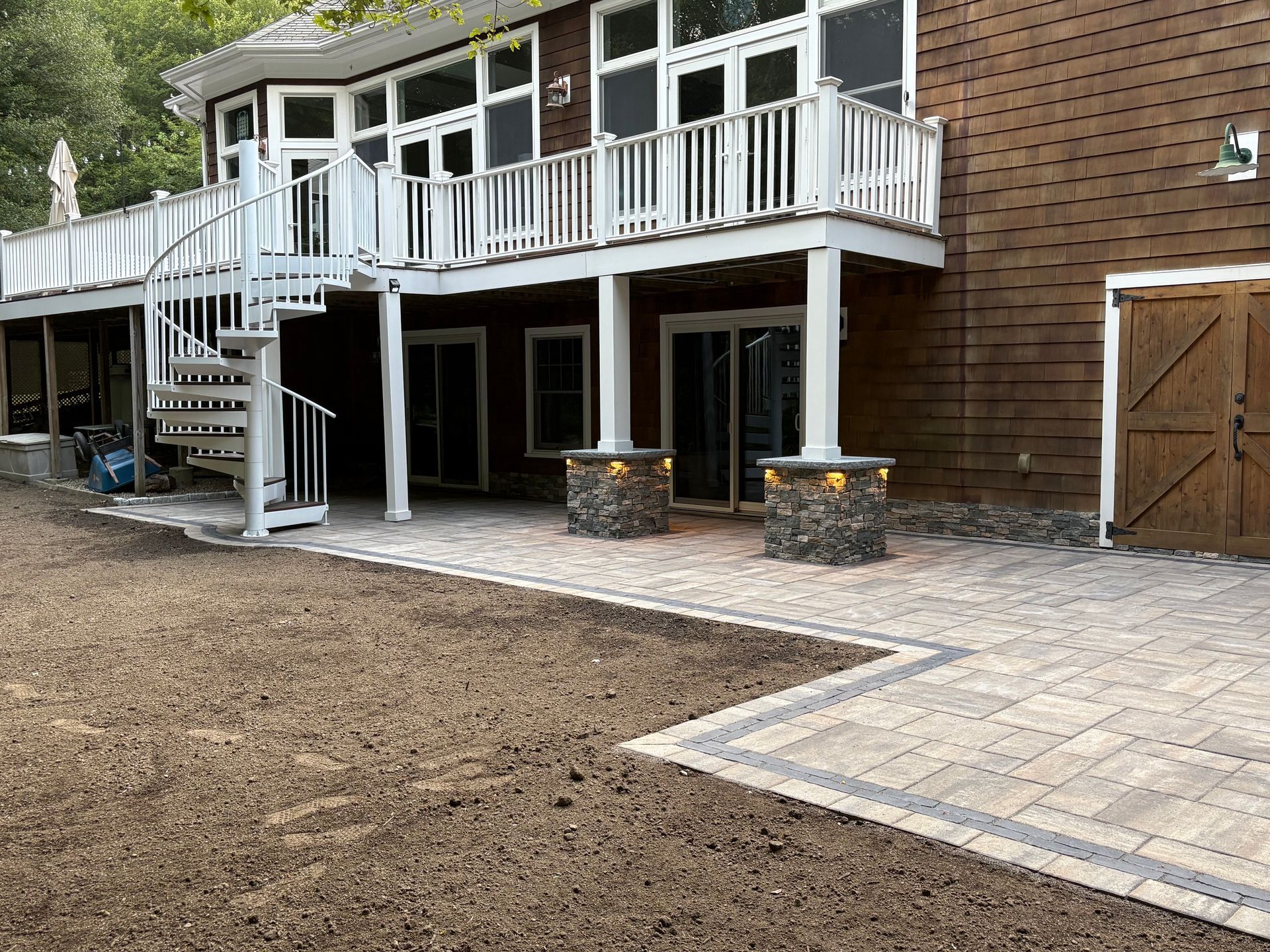 Backyard patio with a white-railed deck, spiral staircase, and stone pillars beside a brick house.