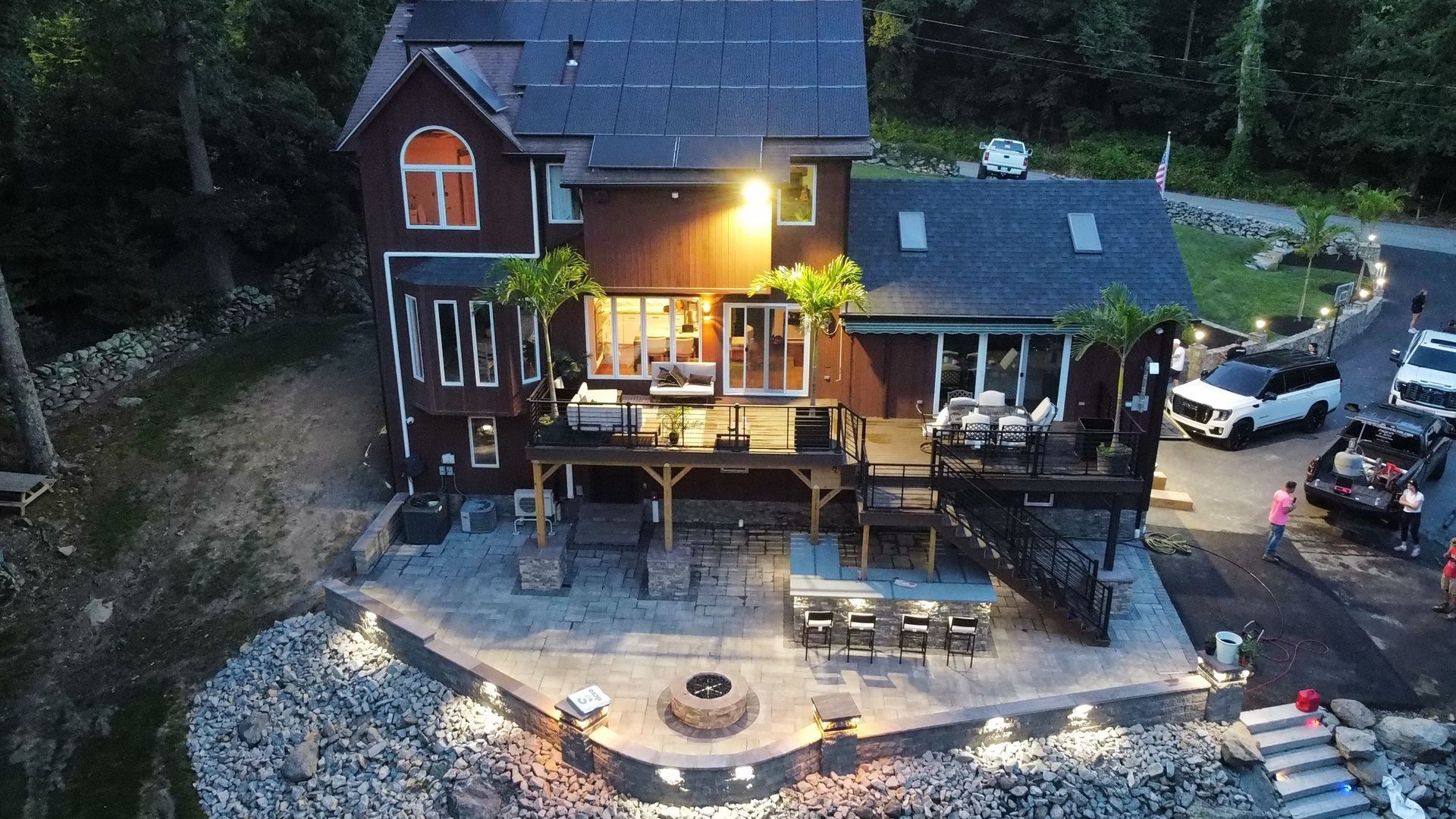 Dusk view of a lit lakeside cabin with decks, patio seating, and parked cars by the water