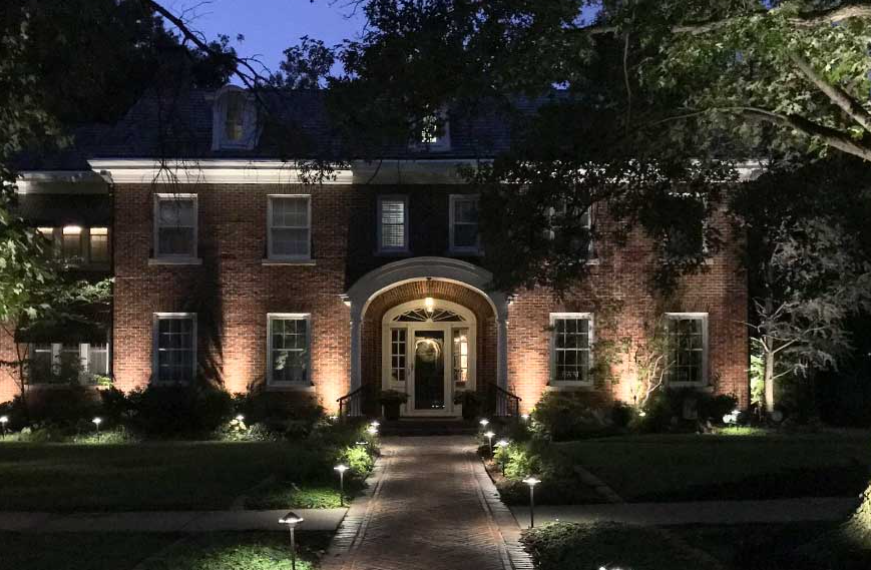 Illuminated brick house at dusk with centered front entrance and lit walkway through landscaped yard