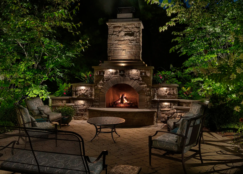Stone outdoor patio with lit fireplace and wicker seating surrounded by trees at night