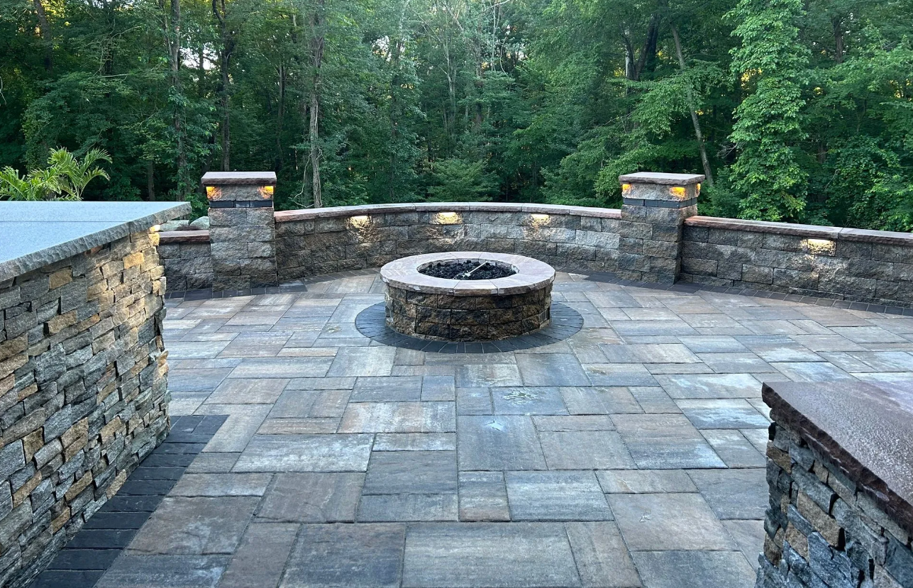 Stone patio with a circular fire pit and low stone walls, overlooking a wooded area.