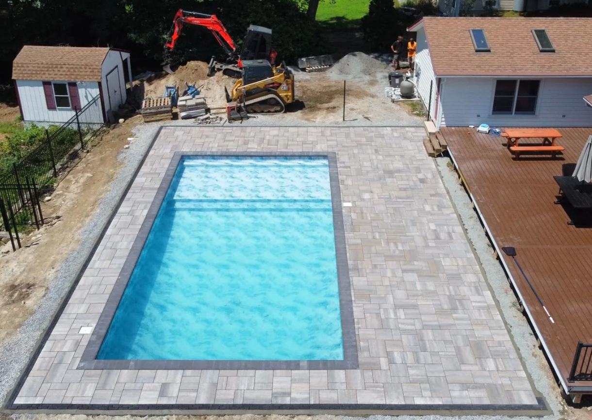 Backyard with rectangular swimming pool, stone patio, wooden deck, and construction equipment nearby