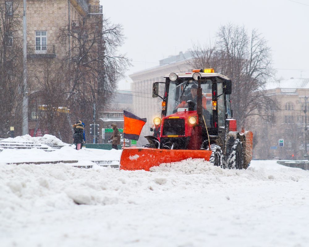 Orange snowplow clearing a snowy street in a city during heavy snowfall